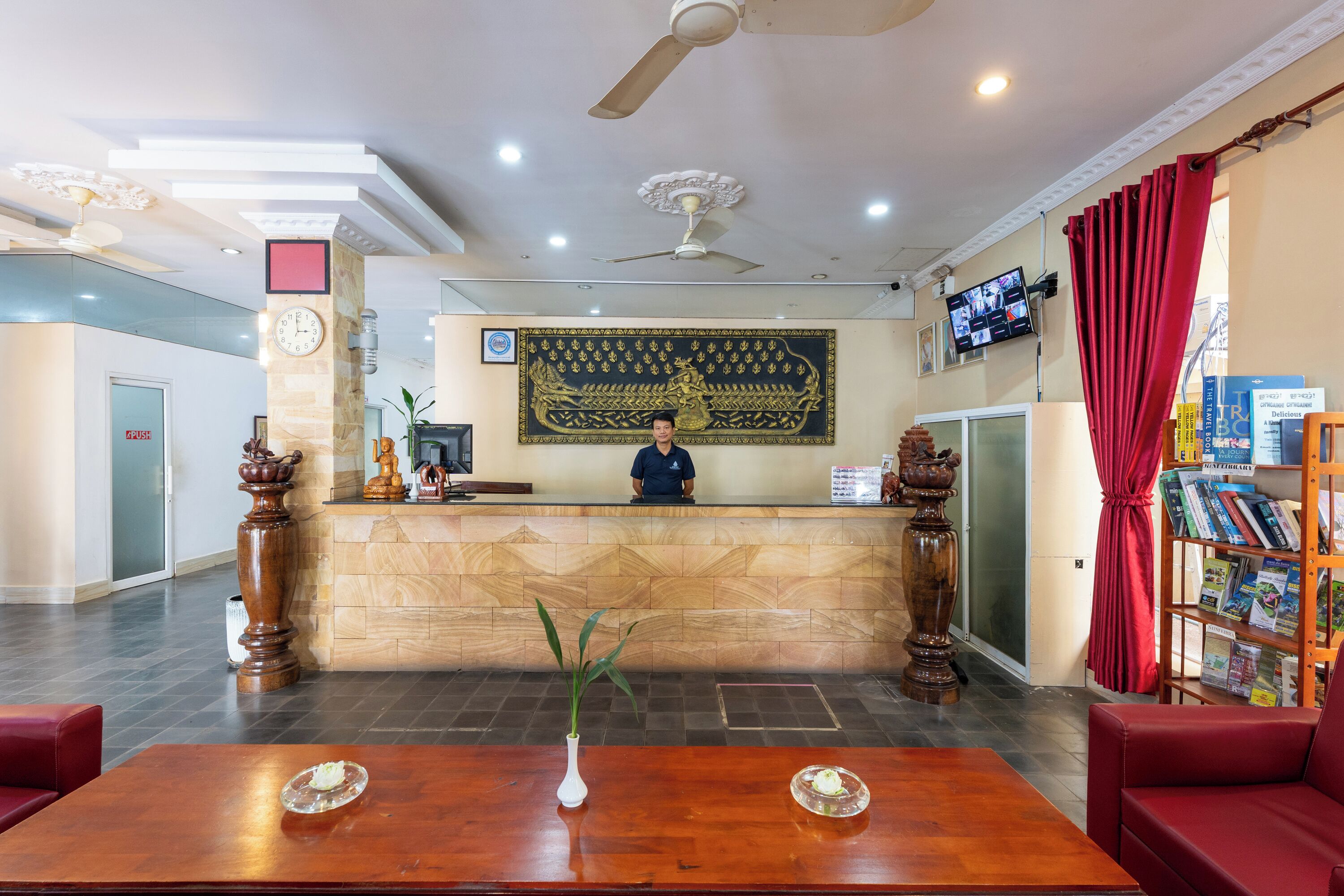 Ein Hotel-Rezeptionsbereich mit Rezeptionist, Empfangstresen, einem großen Wandteppich, einem Fernseher und einer Bücherwand mit Zeitschriften.