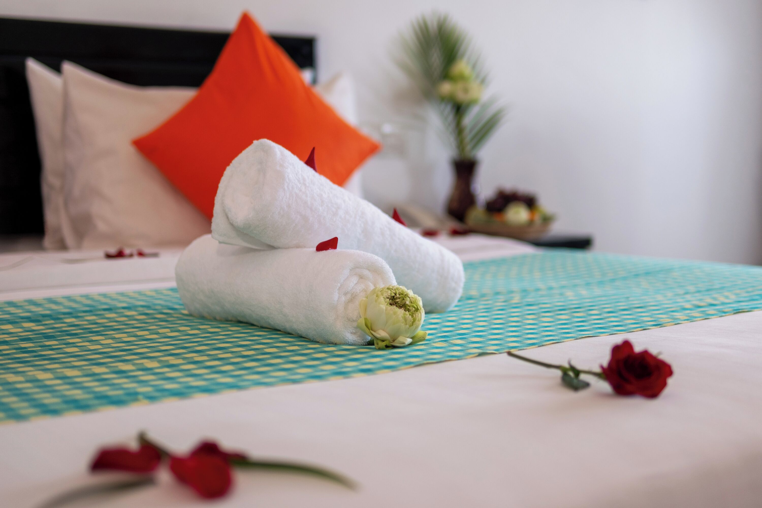 A neatly made bed with white and orange pillows, a green checkered bedspread, and a red rose on the bed.