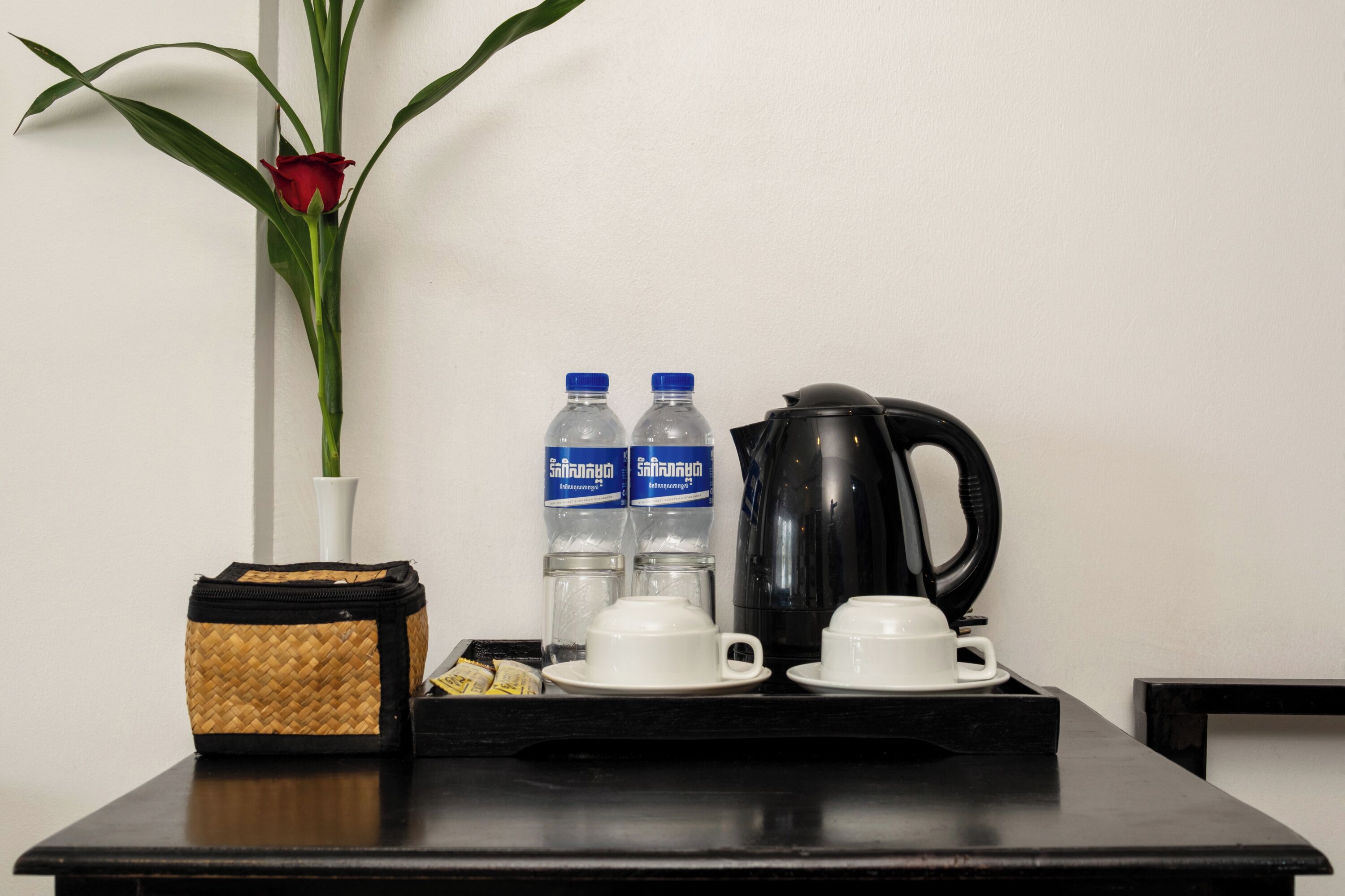 Ein Tisch mit einer Tablettablage, darauf eine weiße Tasse, eine schwarze Kaffeekanne, zwei Wasserflaschen, eine gewebte Tasche und eine rote Rose in einer weißen Vase.
