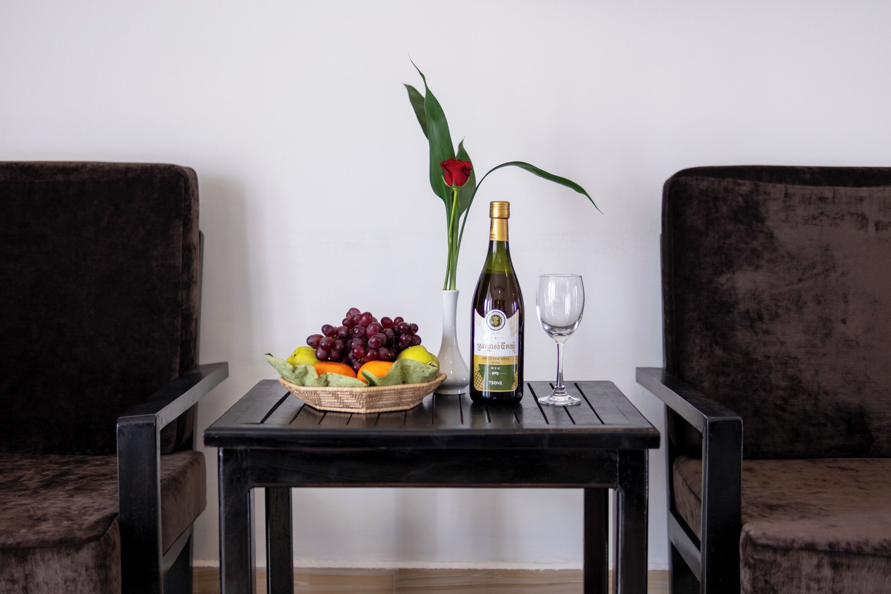 A table with a bottle of wine, a glass, a bowl of fruit, and a vase with a red rose.