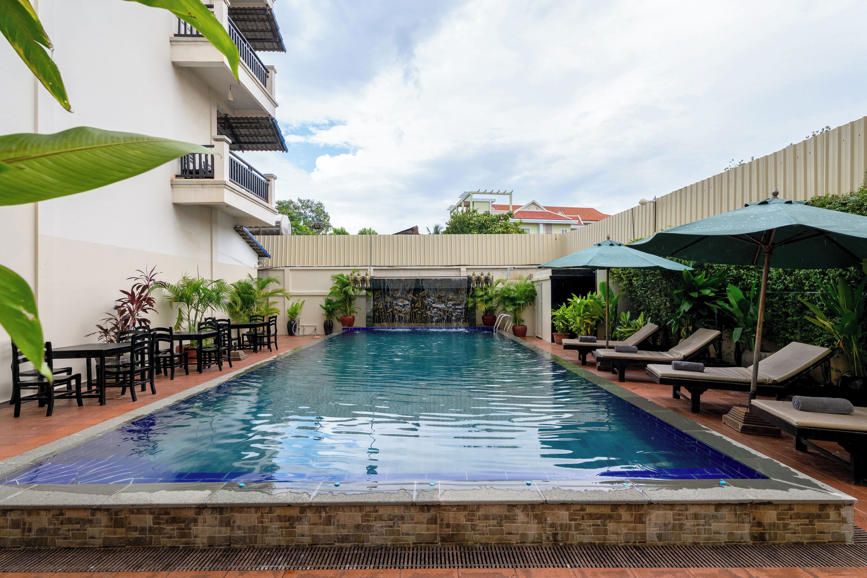 A rectangular swimming pool with a waterfall feature, surrounded by lounge chairs and umbrellas, with a white building and a fence in the background.