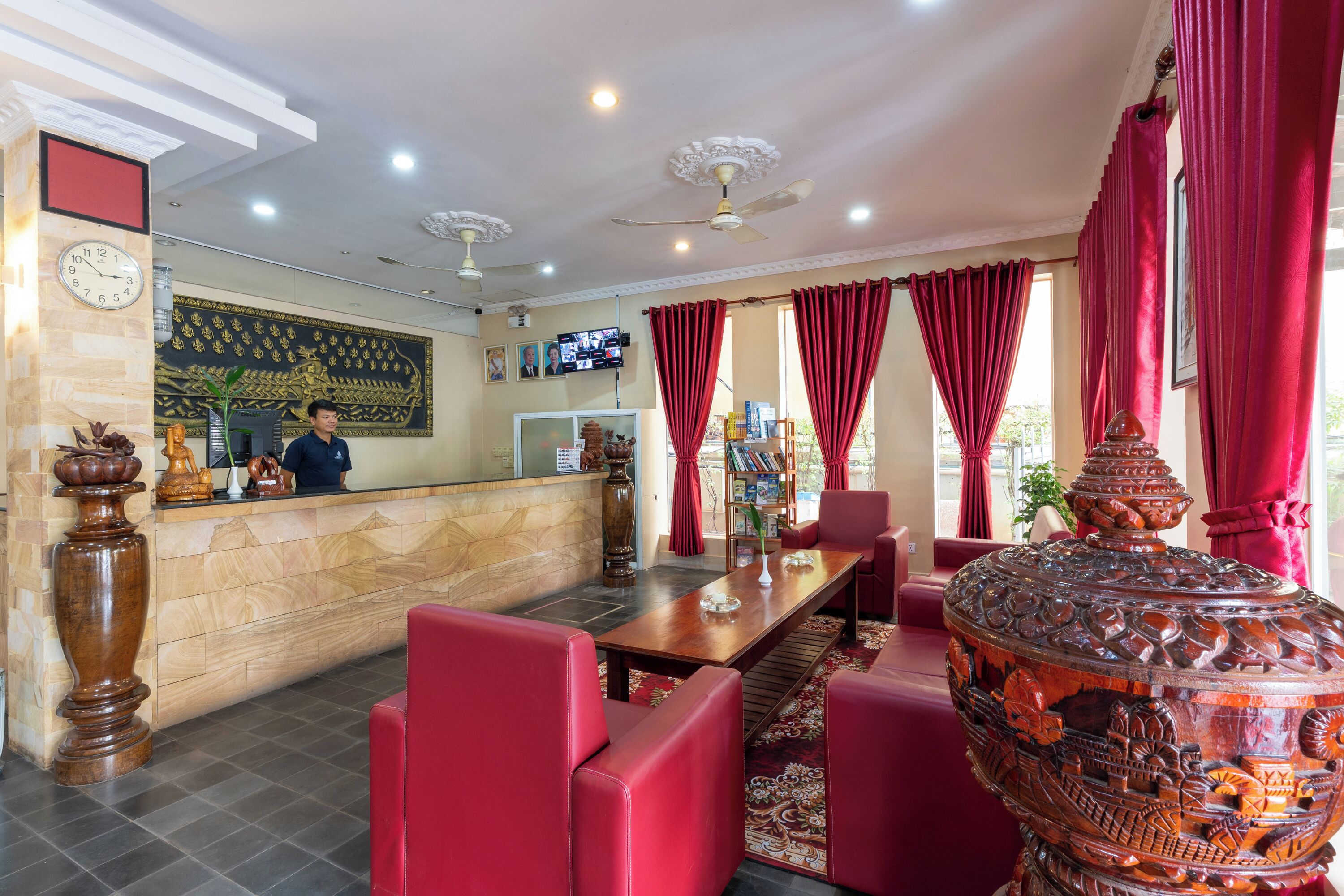 Eine Hotelhalle mit einer Rezeption, roten Polstersitzgelegenheiten, einer großen Dekorvase und einem Fernsehbildschirm, der eine Videokonferenz zeigt.