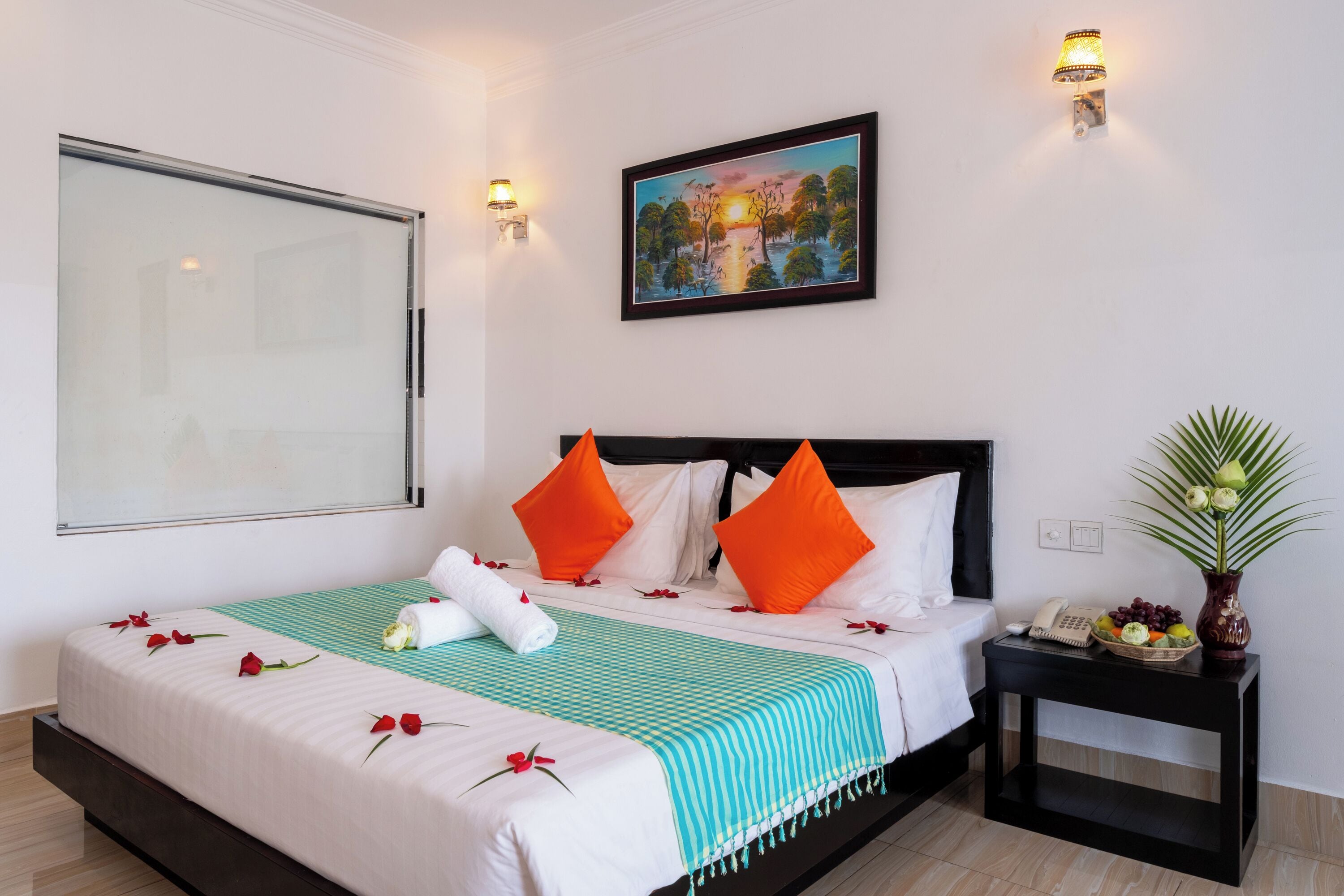 Un lit bien fait, agrémenté d’oreillers blancs et orange, d’une couverture rayée turquoise et blanche, et de pétales de roses rouges disposés sur le lit. Un tableau orne le mur, ainsi qu’une petite table sur laquelle est posé un vase de fleurs et un téléphone fixé au mur.