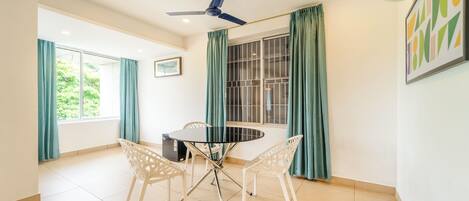 Ein Zimmer mit einem runden Tisch, weißen Stühlen, einem Deckenventilator und einem Fenster mit Vorhängen.