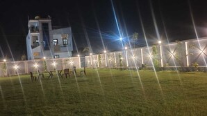 A modern building with a flat roof and multiple windows, surrounded by a well-lit fence and a grassy area with a few chairs. - Hotel The Crystal (Māvli)