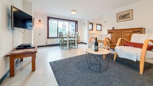 Un salon avec un téléviseur, un coin repas avec des chaises, un meuble en bois et une fenêtre donnant sur un paysage agréable.