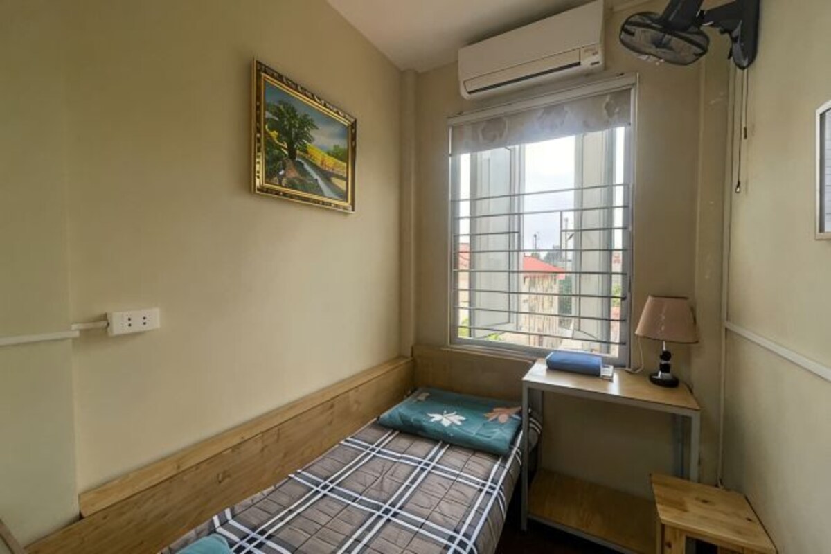 A small room with a bed, a window with bars, a painting on the wall, and a desk with a lamp.
