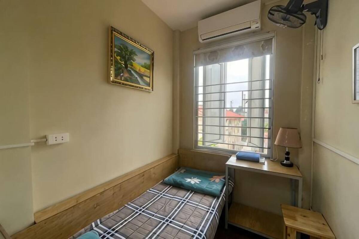 Um quarto pequeno com uma cama, janela com grades, um quadro na parede e uma escrivaninha com abajur.