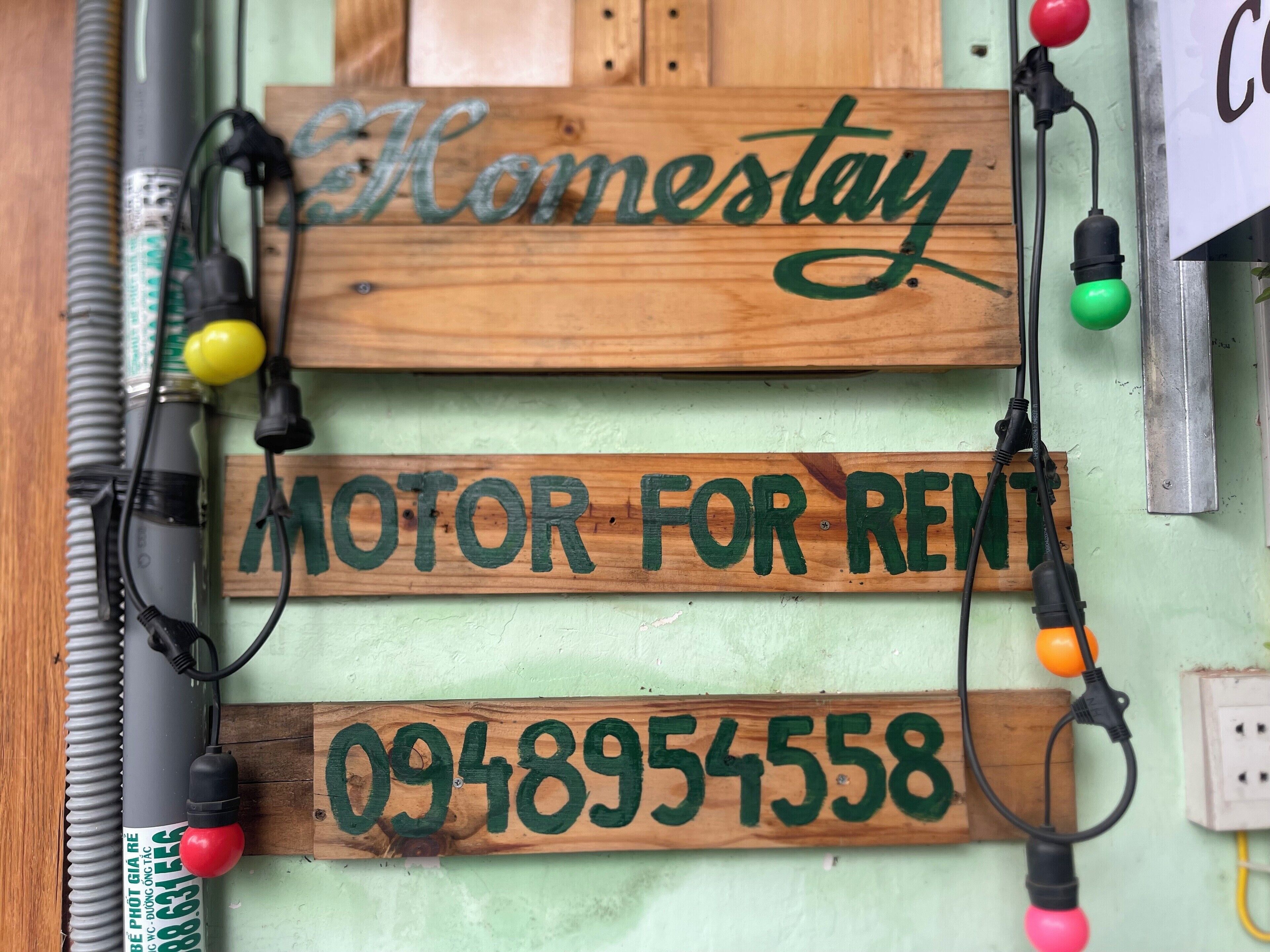 Un letrero de madera con la inscripción 'Homestay' y 'MOTOR POR ALQUILER', junto con un número de teléfono.