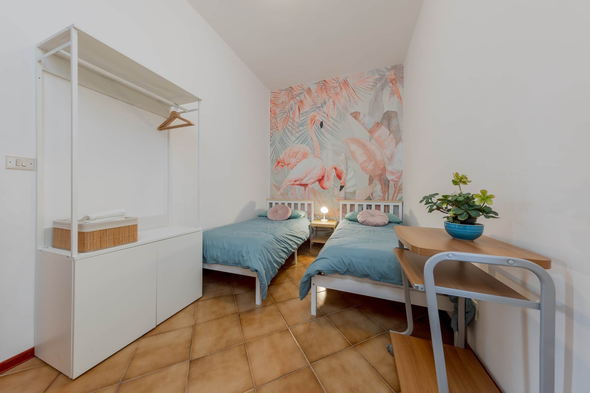 Une petite pièce bien éclairée, avec deux lits simples, une armoire blanche, un bureau en bois et une fresque murale.