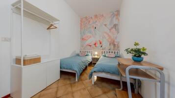 Une petite pièce bien éclairée, avec deux lits simples, une armoire blanche, un bureau en bois et une fresque murale.