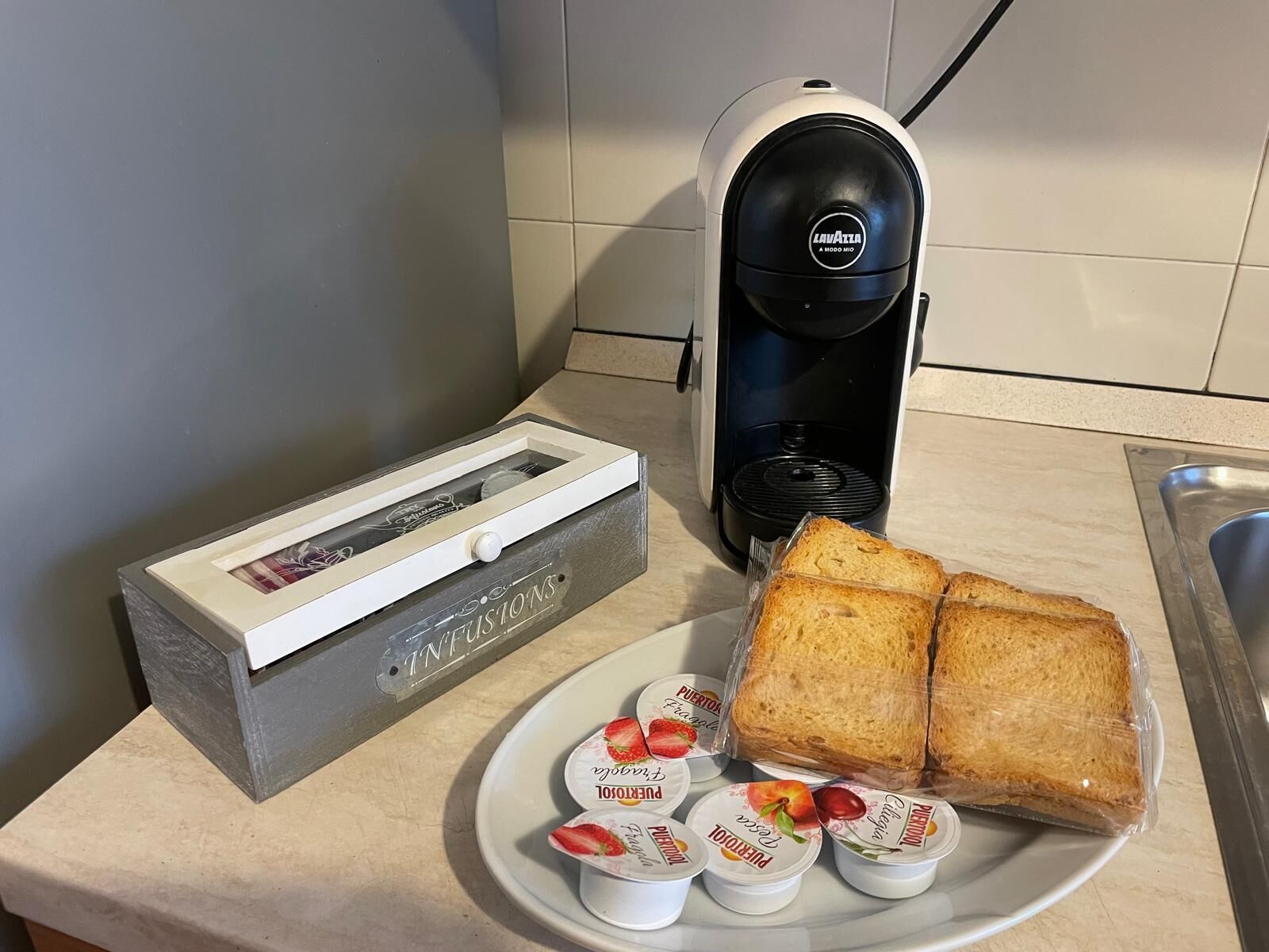 Un plan de travail de cuisine avec une machine à café, un grille-pain, une assiette avec du pain et des pots de yaourt, et une boîte étiquetée « INFUSIONS ».