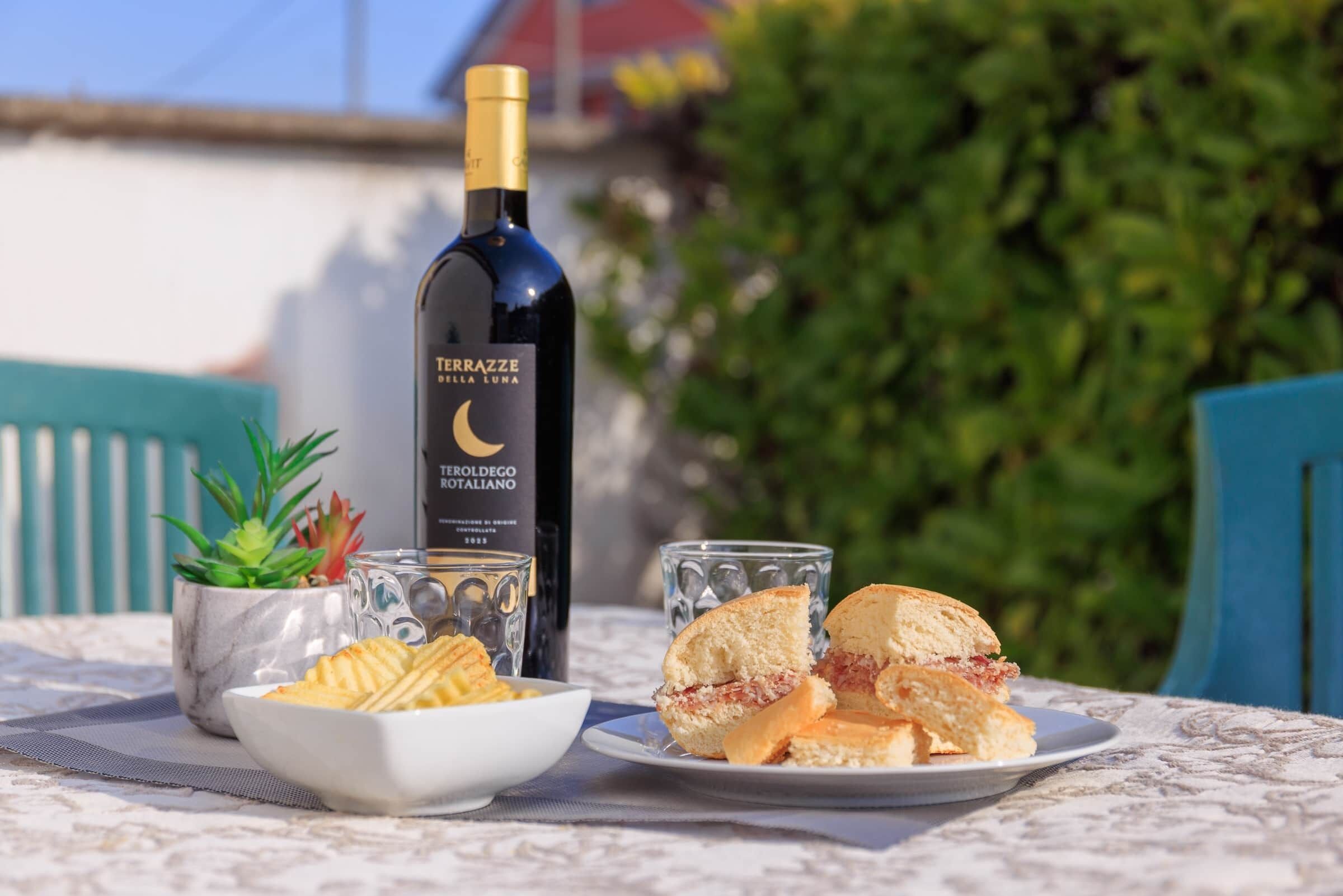 A table with a bottle of Teroldego Rosso, a glass of water, a bowl of crackers, and a sandwich on a tablecloth.