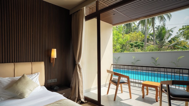Una habitación de hotel con cama, mesita de noche, lámpara y ventana con vista a una piscina y palmeras.