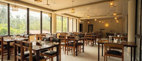 A spacious dining area with wooden tables and chairs, large windows offering a view of greenery, and pendant lighting.