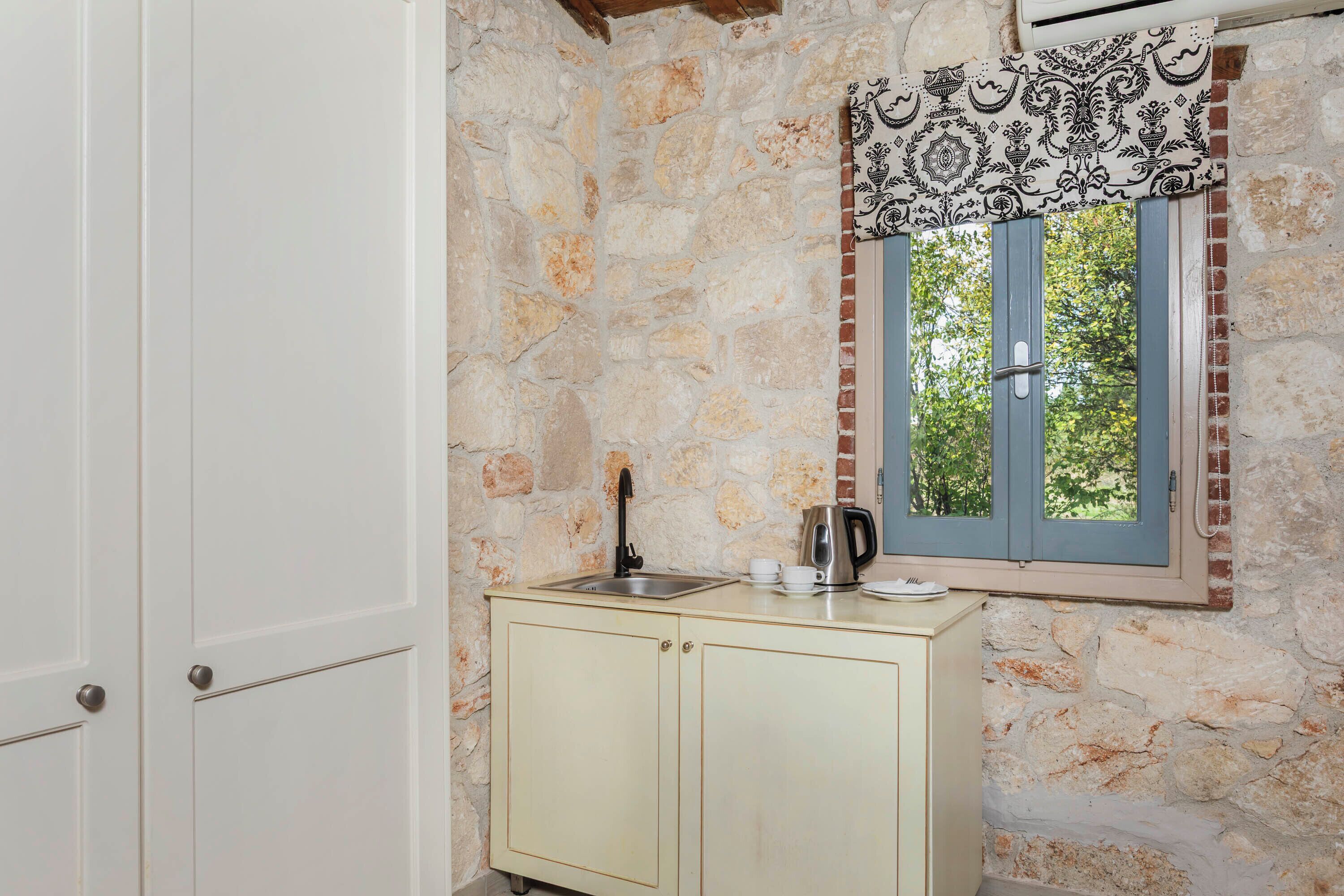 Una cocina con pared de piedra, ventana con contraventanas azules, un lavamanos y un hervidor.