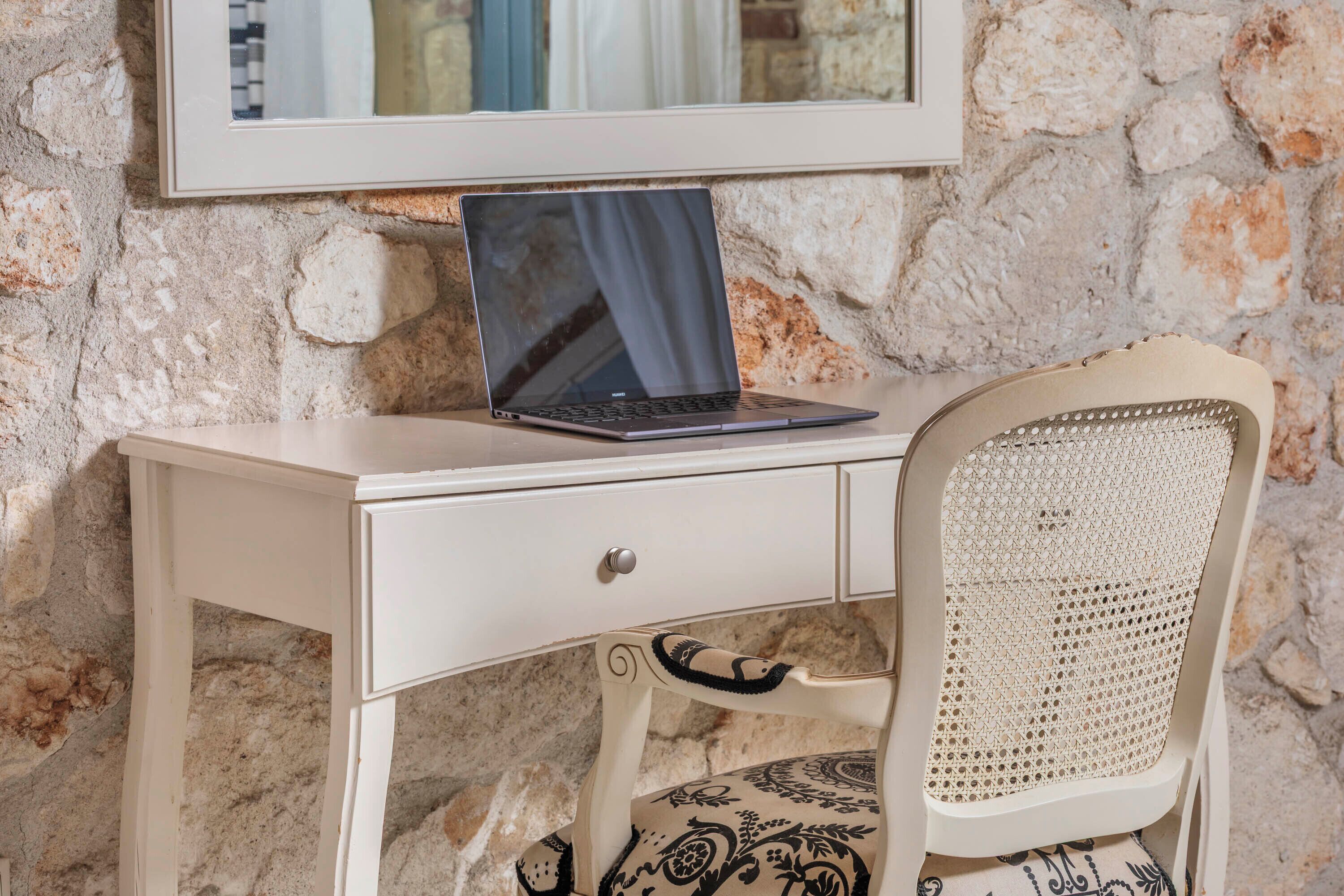 A laptop on a white desk with a drawer, a wicker chair, and a stone wall background.