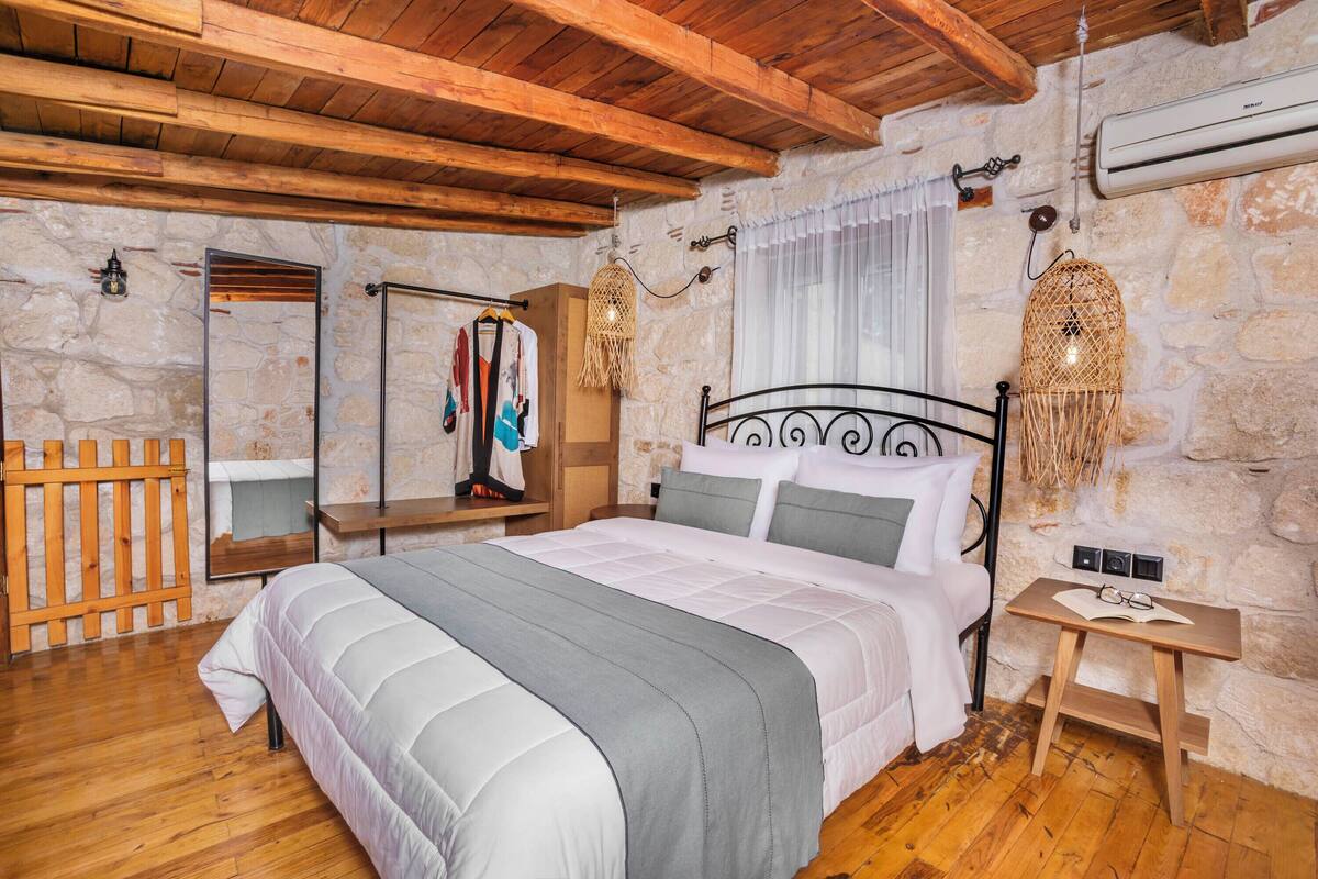 A bedroom with a large bed, wooden beams, stone walls, a wardrobe, and a bedside table with a lamp.