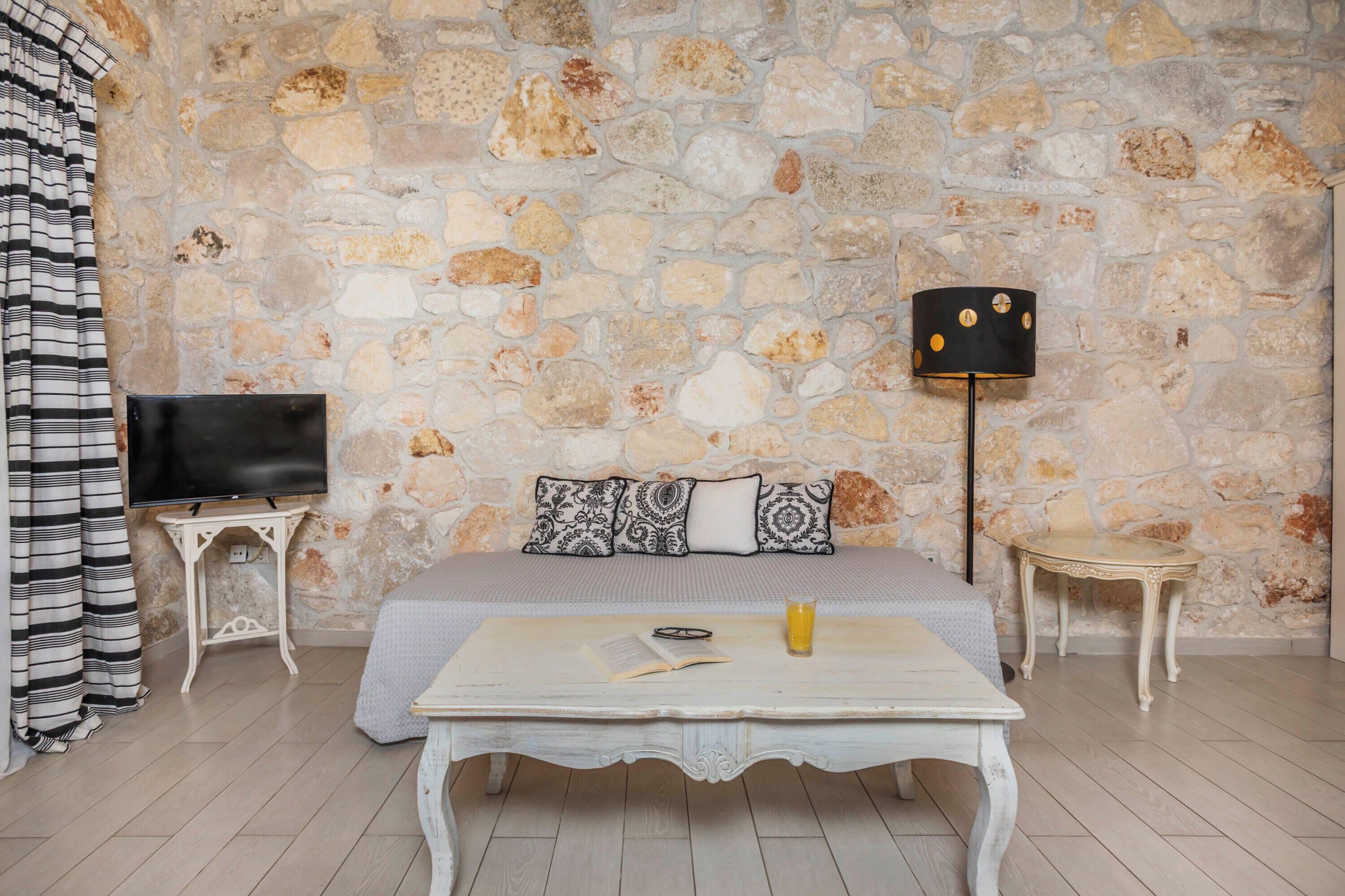 A bedroom with a stone wall, a bed, a TV, a lamp, a table with a book and a glass of orange juice, and a striped curtain.