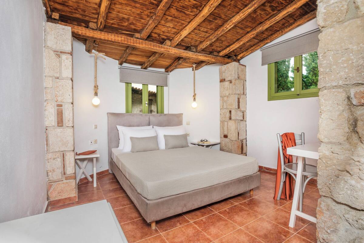 A bedroom with a large bed, stone walls, a wooden ceiling, and a window with a view.