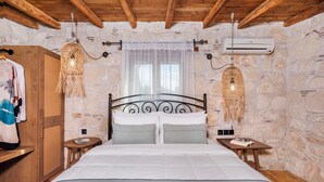 A bedroom with a stone wall, wooden ceiling beams, a bed with white bedding, two bedside tables, and a hanging light fixture. - Petrino Eco Village (Kassandra)