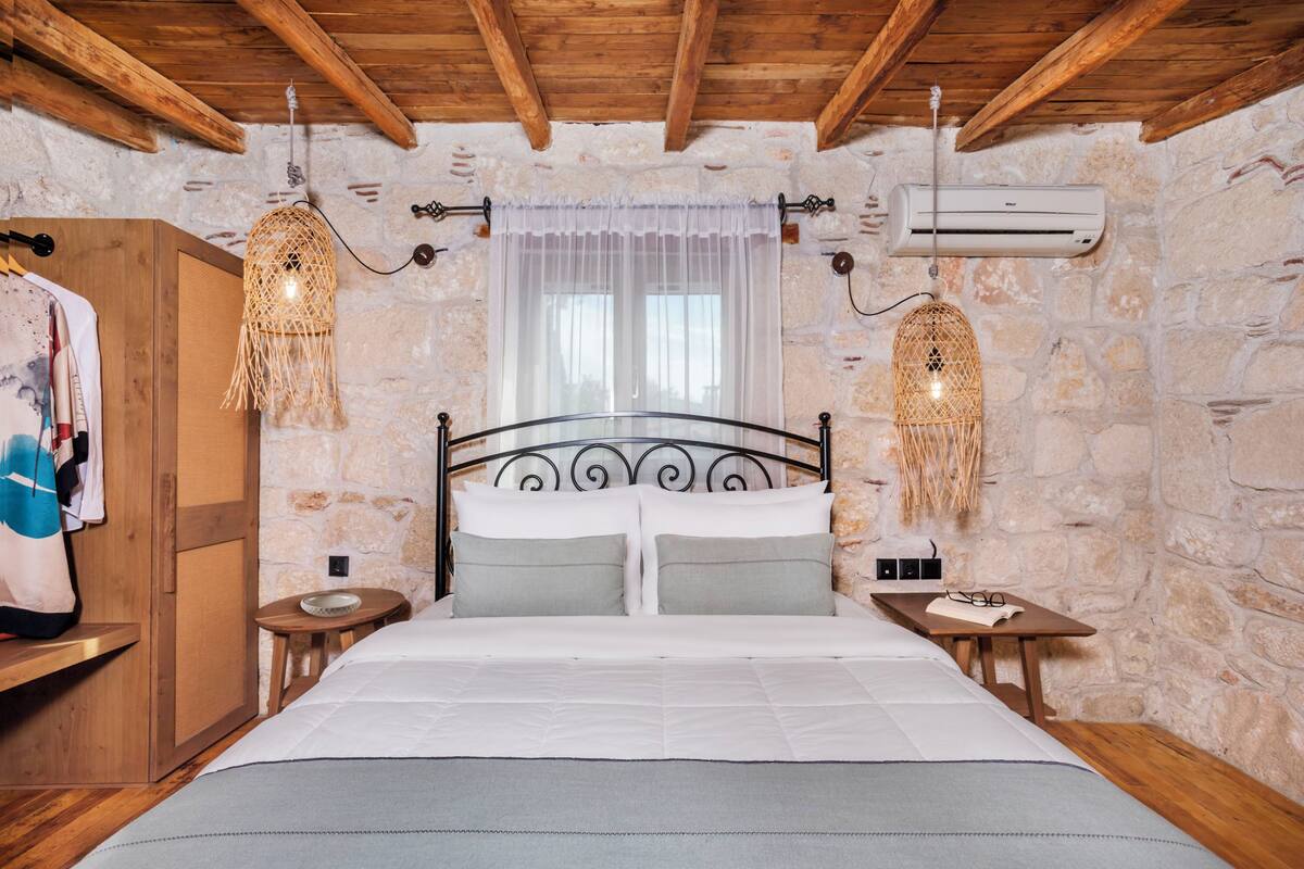 A bedroom with a stone wall, wooden ceiling beams, a bed with white bedding, two bedside tables, and a hanging light fixture.