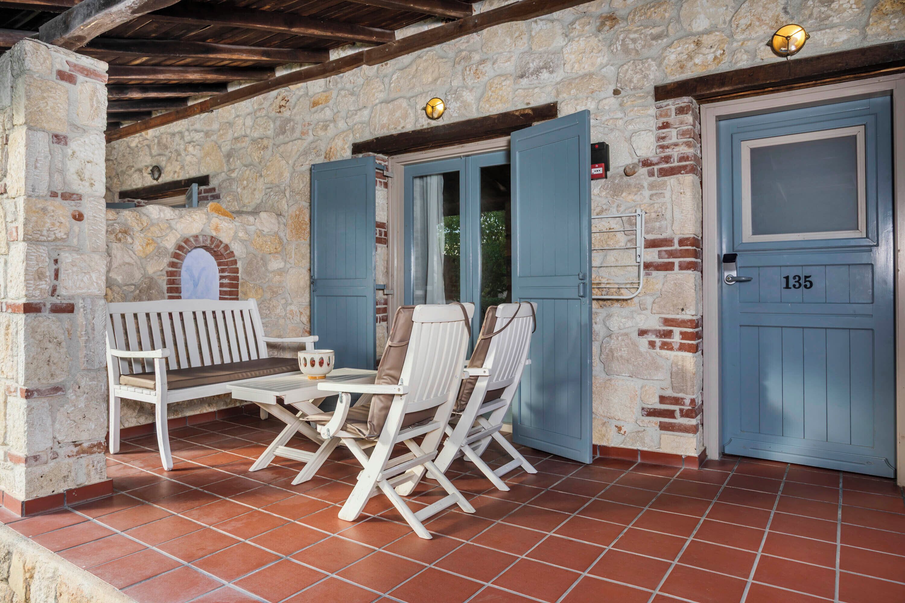 A stone-walled patio with a wooden bench and two white chairs, a tiled floor, and a blue door with the number 135.