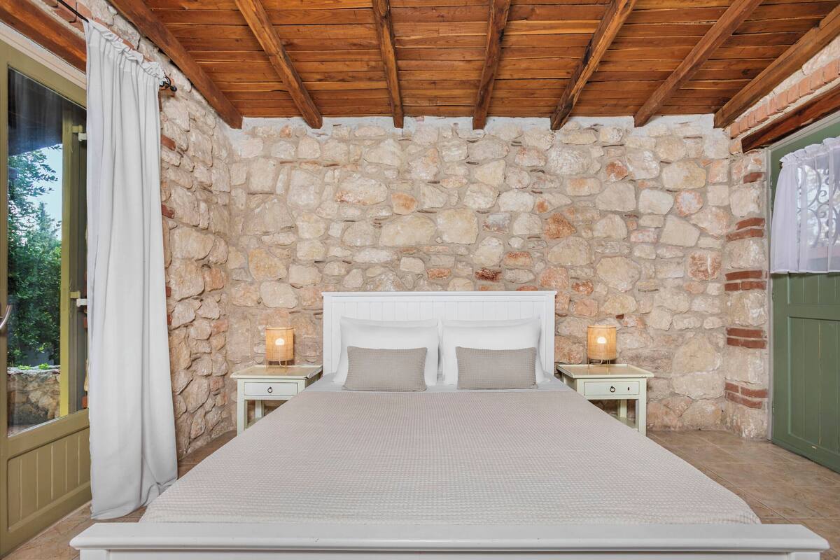 A bedroom with a stone wall, wooden ceiling, a bed, nightstands, and a window with curtains.