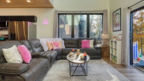 A modern living room with a sectional sofa, a coffee table, and a view of the outdoors through large windows. - Smoky Mountain Cabin with Indoor Pool + Hot Tub + Views | Dolly's Dreamhouse (Sevierville)