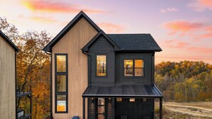 A modern two-story house with a wooden exterior and a black roof, featuring a deck with outdoor seating and a visible sign reading "HOWDY". - Smoky Mountain Cabin with Indoor Pool + Hot Tub + Views | Dolly's Dreamhouse (Sevierville)