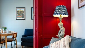 Ein Wohnzimmer mit roter Tür, blauem Sofa, einem Esstisch mit Stühlen und einer Lampe mit gemustertem Lampenschirm.