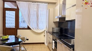 A compact kitchen with a dining table, chairs, a microwave, oven, and a window with sheer curtains. - 2-Bedroom Apartment by Sweet Home (Yerevan)