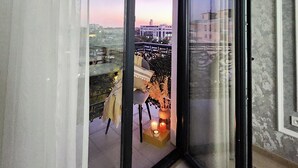 Une pièce dotée d’une grande porte-fenêtre coulissante donnant sur un balcon extérieur, d’où l’on peut admirer le panorama de la ville au coucher du soleil.
