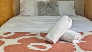 Un lit bien fait, avec du linge de lit blanc, un oreiller moelleux de couleur grise et une serviette blanche enroulée, posés sur un tapis orange à motifs.