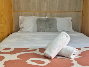 A neatly made bed with white linens, a fluffy grey pillow, and a rolled white towel on a patterned orange carpet. - Modern 4 Bedrooms Home | Free Parking | Walk to Station (Sefton)