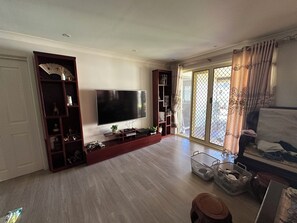 A living room with a large flat-screen TV, wooden entertainment unit, and a sliding glass door leading to a balcony. - Modern 4 Bedrooms Home | Free Parking | Walk to Station (Sefton)