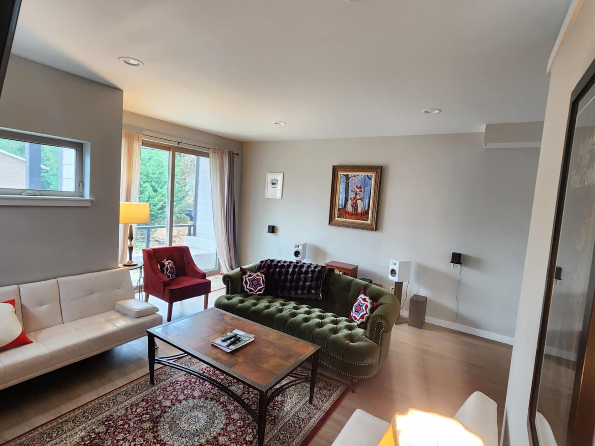 A modern living room with a green sofa, a red armchair, and a white sofa. There is a coffee table in the center, a patterned rug on the floor, and a painting on the wall.