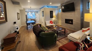 Una sala de estar moderna con sofá, mesa de centro y televisión. Se observa una chimenea y un comedor al fondo.