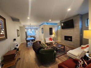 A modern living room with a sofa, coffee table, and TV. There is a fireplace and a dining area in the background. - Three-story Victorian-ish Rocker Townhome in St. John’s! (Portland)
