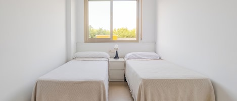 Una habitación pequeña y limpia con dos camas individuales, una mesita de noche con lámpara y una ventana con vistas al exterior.