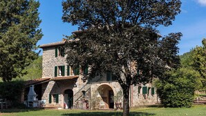 Un edificio de piedra con un gran árbol en frente, un espacio de estar al aire libre techado y una escalera de piedra que conduce a la entrada.