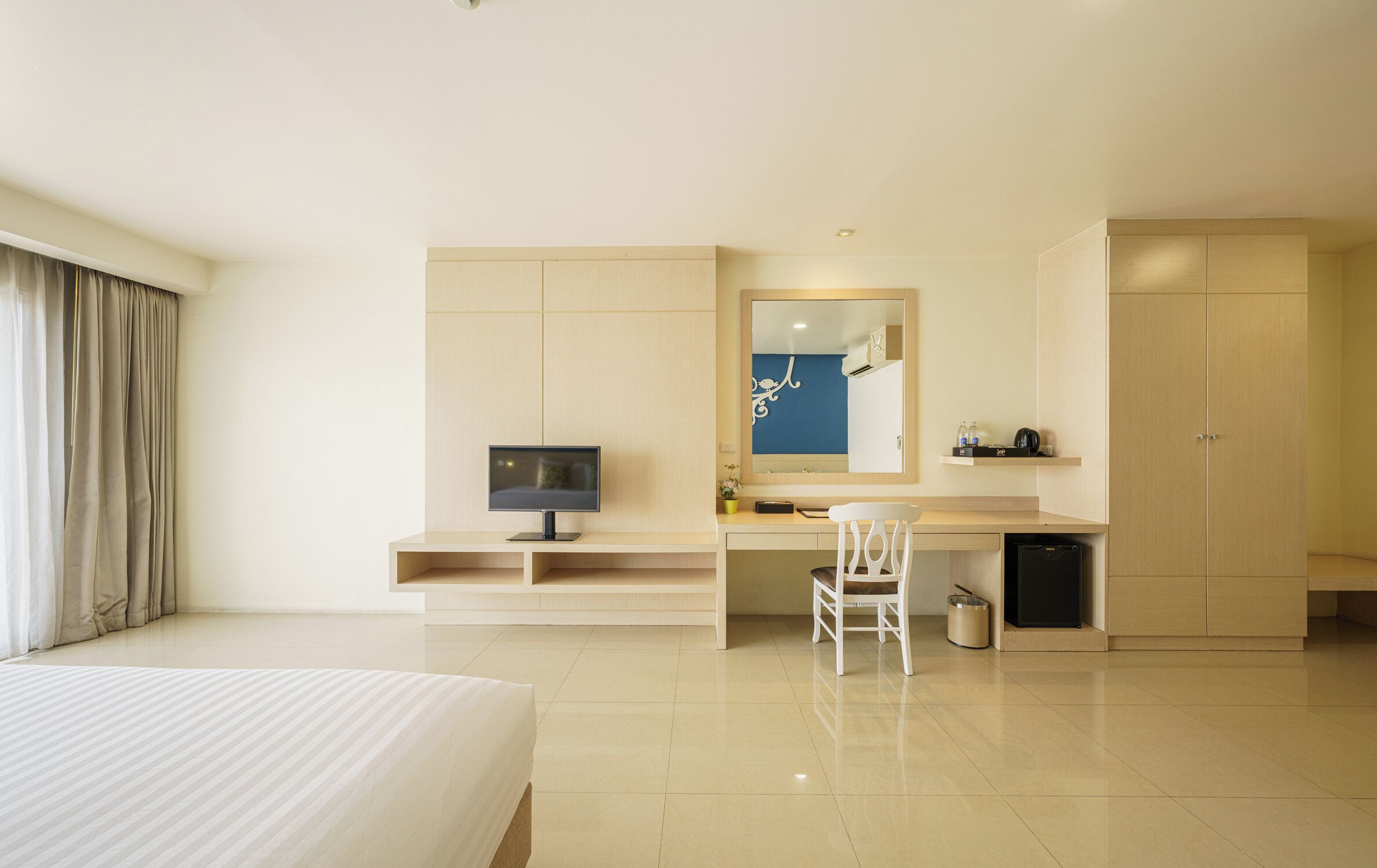 A hotel room with a bed, a desk with a computer, a chair, a wardrobe, and a mirror.
