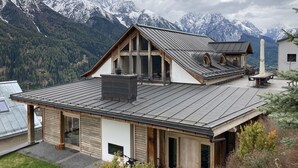 Un moderno chalet con tetto spiovente, ampie finestre e un camino, sullo sfondo di montagne innevate.