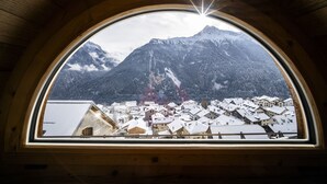 Vista attraverso una finestra con cornice in legno su un borgo montano con edifici imbiancati dalla neve e una montagna imponente sullo sfondo.