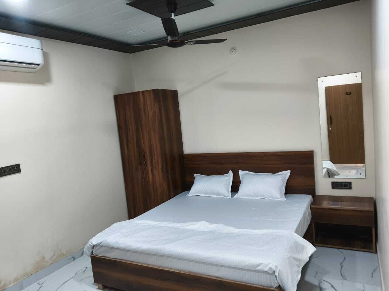 A bedroom with a wooden bed frame, white bedding, a ceiling fan, an air conditioning unit, a wooden wardrobe, and a bedside table with a mirror.