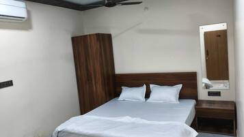 A bedroom with a wooden bed frame, white bedding, a ceiling fan, an air conditioning unit, a wooden wardrobe, and a bedside table with a mirror.