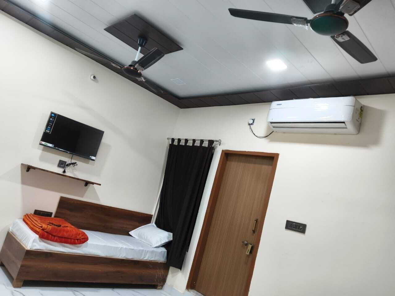 A bedroom with a bed, a ceiling fan, an air conditioner, a TV mounted on the wall, and a door.