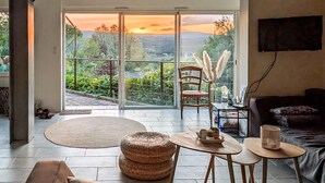 Un salon comprenant un canapé, une table basse, un fauteuil et une vue sur un coucher de soleil, visible à travers une porte vitrée.