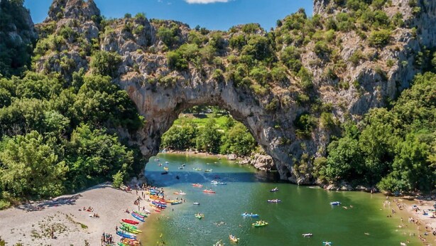 Une arche naturelle en pierre surplombe une rivière où l’on voit des bateaux et des personnes sur la rive.