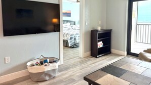 Una sala de estar con un televisor de pantalla plana montado en la pared, un mueble de entretenimiento negro, una cesta de mimbre y vistas al balcón a través de una puerta abierta.